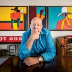 Frank Palmer Sitting At Desk With One Hand On Chin And Paintings Behind him