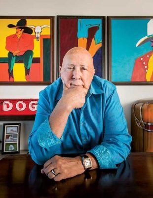 Frank Palmer Sitting At Desk With One Hand On Chin And Paintings Behind him