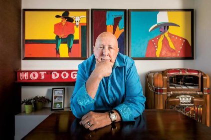 Frank Palmer Sitting At Desk With One Hand On Chin And Paintings Behind him