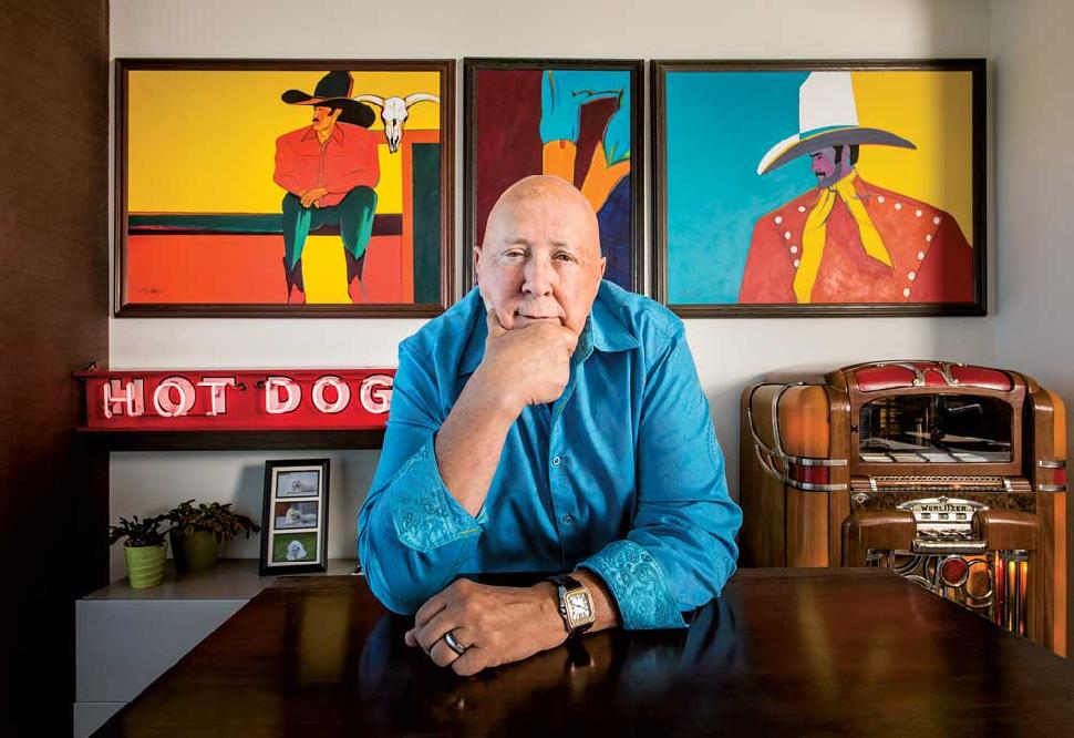 Frank Palmer Sitting At Desk With One Hand On Chin And Paintings Behind him