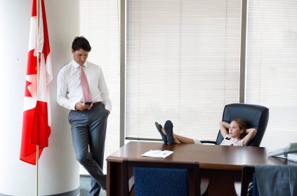 Justin Trudeau Standing Looking At Mobile Phone With Daughter Sitting At his Desk With Feet On Table