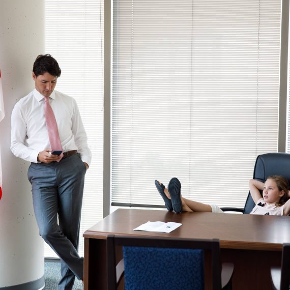 Justin Trudeau Standing Looking At Mobile Phone With Daughter Sitting At his Desk With Feet On Table