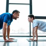 Grown Man And Teenage Boy Looking At Each Other While Doing Push-Ups