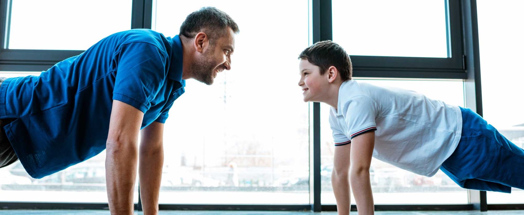 Grown Man And Teenage Boy Looking At Each Other While Doing Push-Ups