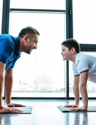 Grown Man And Teenage Boy Looking At Each Other While Doing Push-Ups