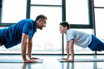 Grown Man And Teenage Boy Looking At Each Other While Doing Push-Ups