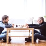 Young Man And Older Man Facing Each Other Sitting Down Playing Check