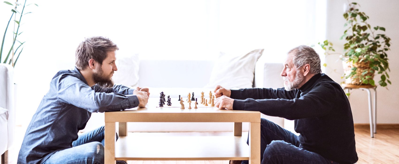 Young Man And Older Man Facing Each Other Sitting Down Playing Check