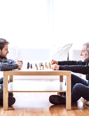 Young Man And Older Man Facing Each Other Sitting Down Playing Check