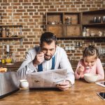 Father Reading Newspaper While Eating Bowl Of Cereal With Daughter