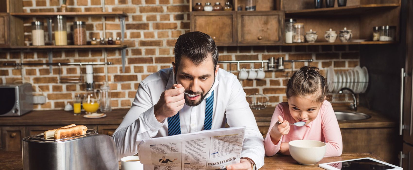 Father Reading Newspaper While Eating Bowl Of Cereal With Daughter
