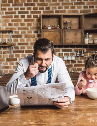 Father Reading Newspaper While Eating Bowl Of Cereal With Daughter