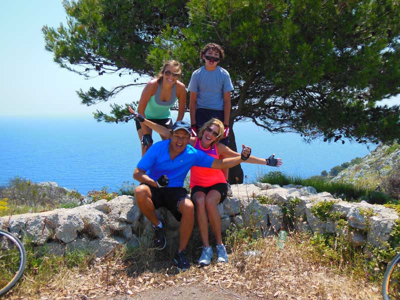 Oscar Munoz With Family Sitting On Rock Fence Overlooking Ocean Smiling And Waving Cycling