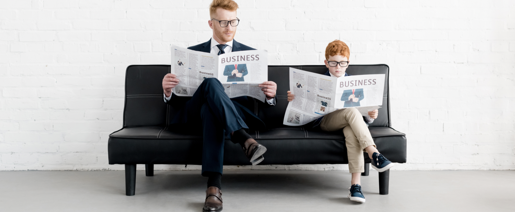 Man And Young Boy Sitting On Sofa Each Reading A Business Newspaper