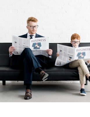 Man And Young Boy Sitting On Sofa Each Reading A Business Newspaper