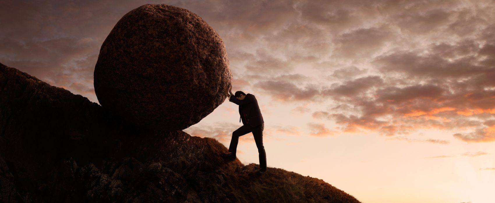 Man Pushing Large Boulder Up A Hill At Dusk