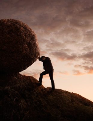 Man Pushing Large Boulder Up A Hill At Dusk