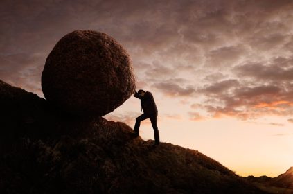 Man Pushing Large Boulder Up A Hill At Dusk
