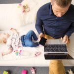 Man Sitting On Sofa Working On Laptop With Sleeping Child Beside Him