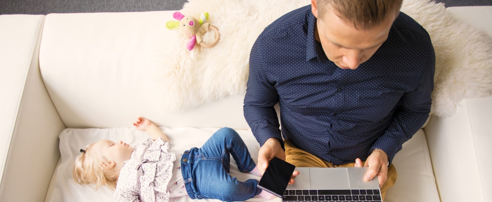 Man Sitting On Sofa Working On Laptop With Sleeping Child Beside Him