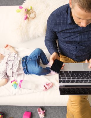 Man Sitting On Sofa Working On Laptop With Sleeping Child Beside Him