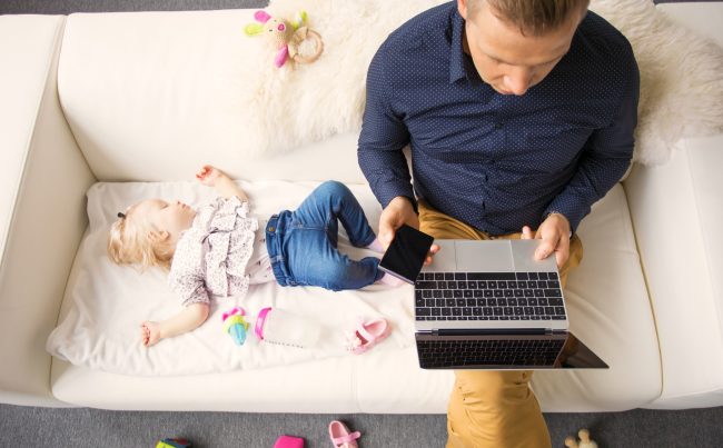 Man Sitting On Sofa Working On Laptop With Sleeping Child Beside Him